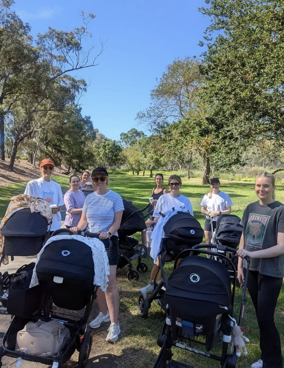 Group of women with strollers standing on a park path with trees and grass under a clear blue sky.