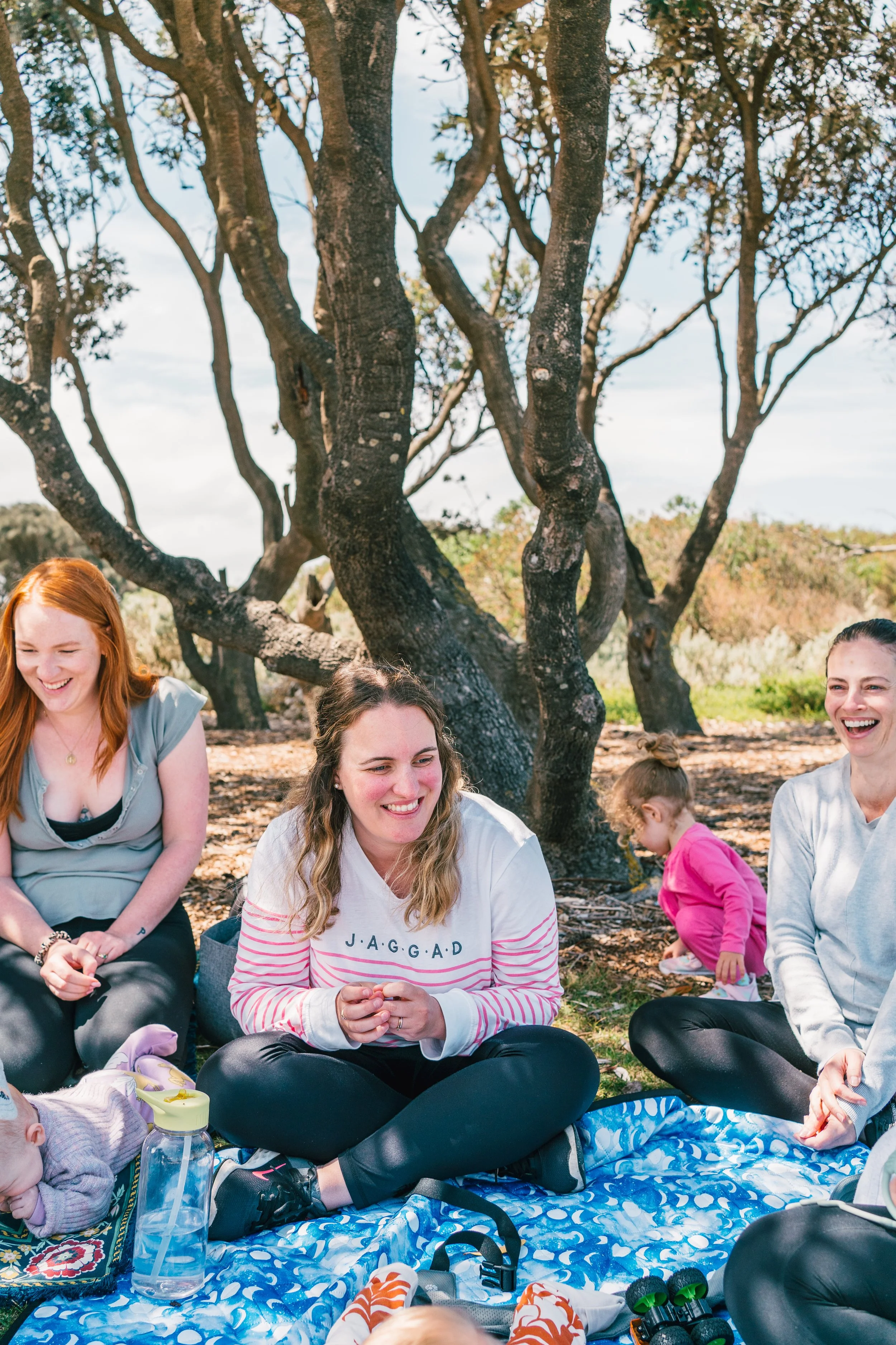 A group of women and children sitting on a blue patterned blanket outdoors under a large tree, smiling and enjoying a picnic on a sunny day.