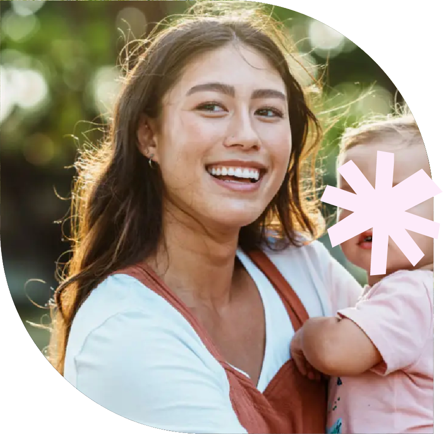 A woman smiling outdoors while holding a young child, with sunlight and blurred green foliage in the background.