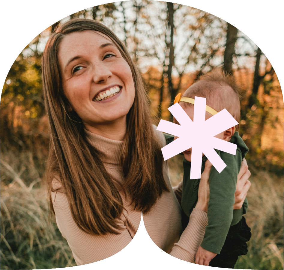 A woman holding a small child outdoors during autumn, with trees in the background and warm lighting.