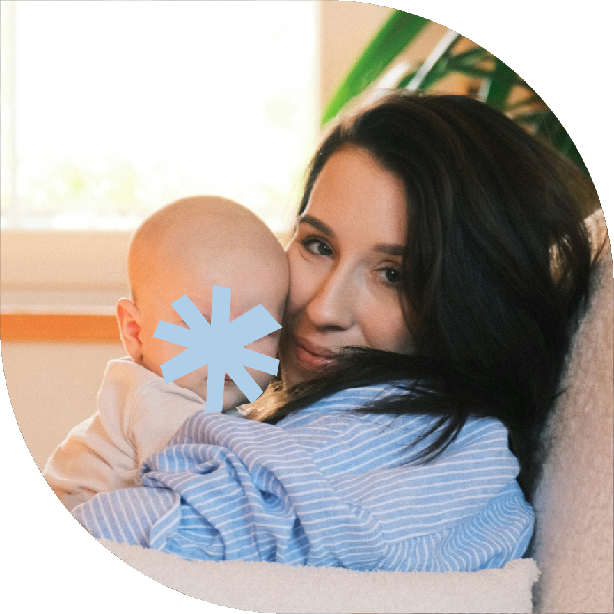 A woman with long dark hair cuddling a baby, both indoors near a window with sunlight and green plants in the background.