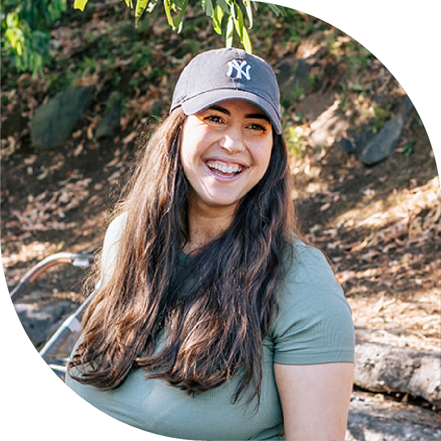 Smiling woman with long brown hair wearing a gray New York Yankees baseball cap and a green t-shirt outdoors.