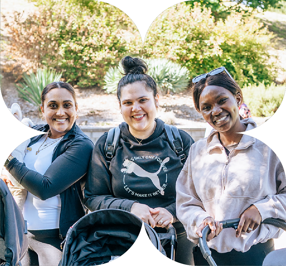 Three women smiling outdoors with greenery in the background. They are standing close together, with one woman holding a stroller.