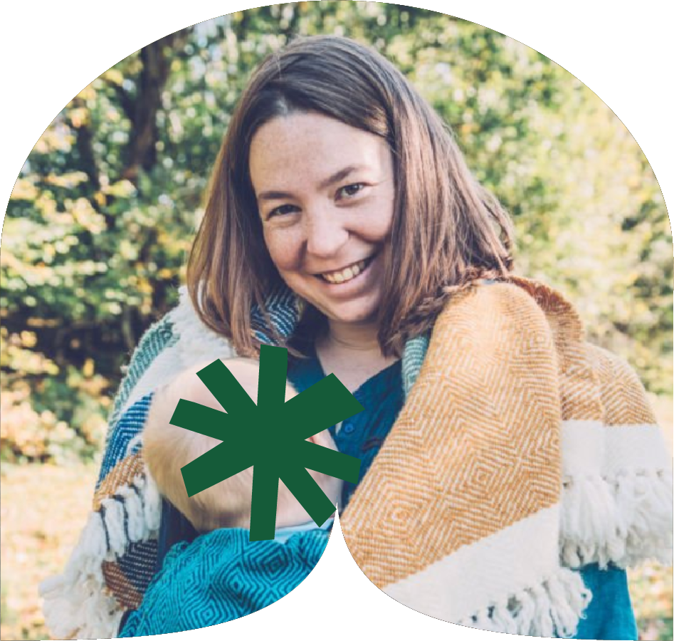 A woman smiling outdoors, wearing a colorful blanket over her shoulders, with trees and sunlight in the background.