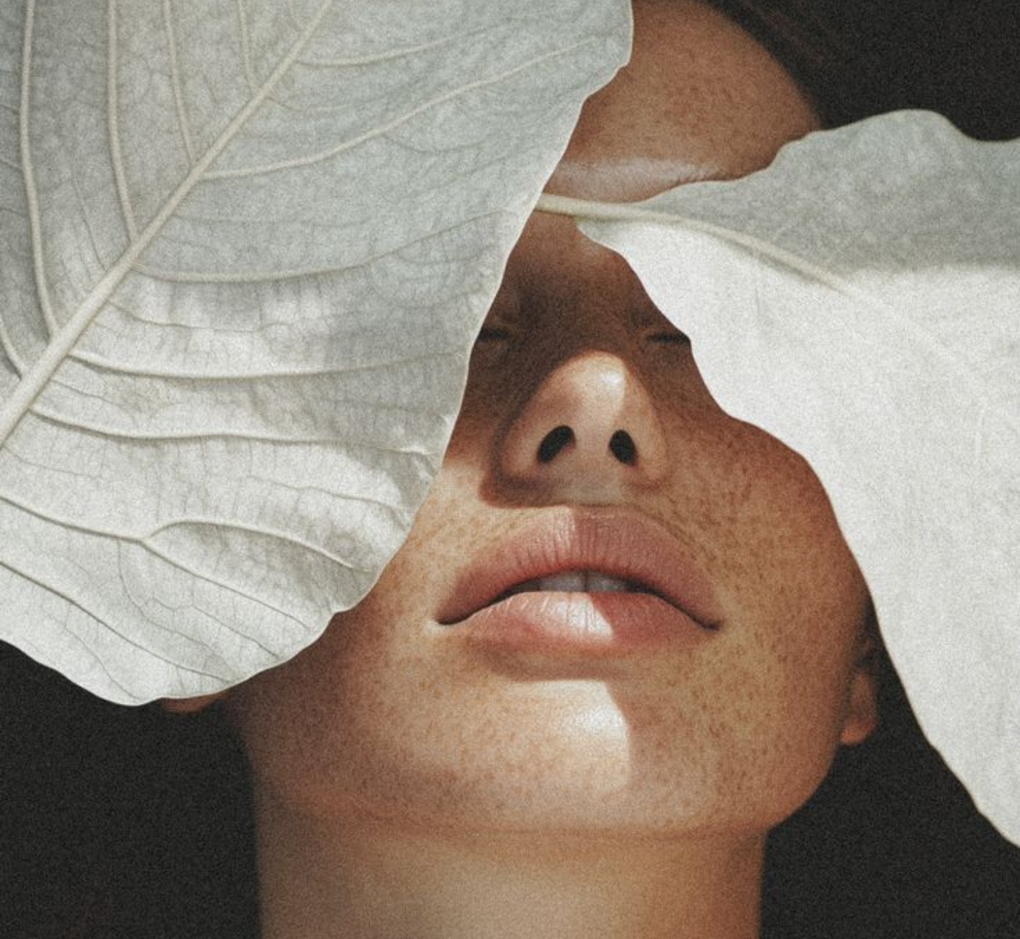Close-up of a woman's face with freckles, partially covered by large white leaves, showing her nose and lips.