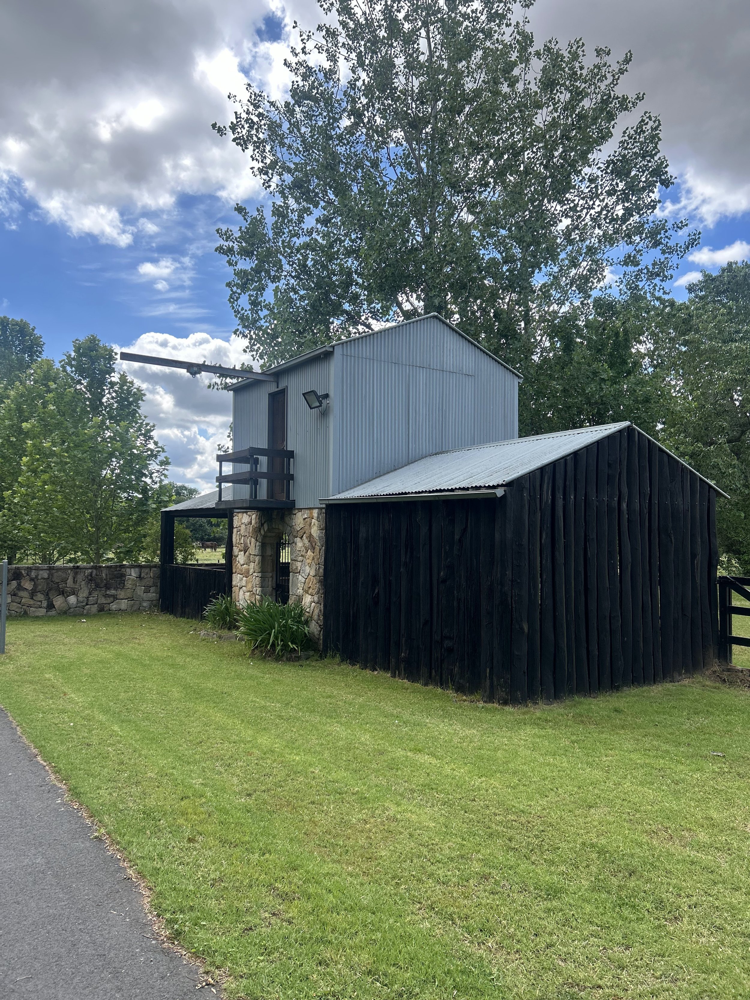 A two-story building with a stone base, black wooden siding, and a metal upper structure, situated on a grassy lawn with trees and a cloudy sky in the background.