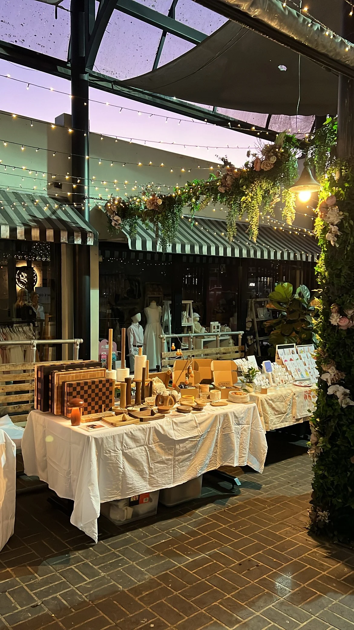 Outdoor market stand with tables displaying various handcrafted items, set under string lights and decorated with green foliage and flowers, at dusk.