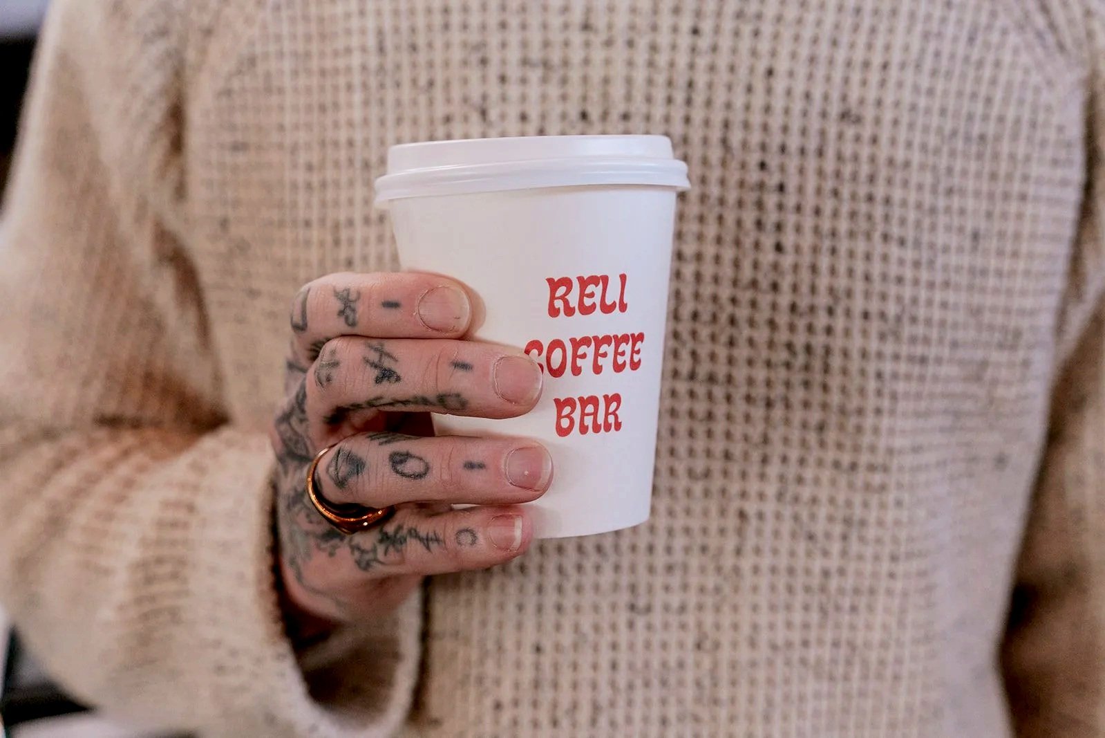 Person holding a white coffee cup with red text that reads 'Rei Coffee Bar', wearing a beige textured sweater, with visible tattoos on their fingers.