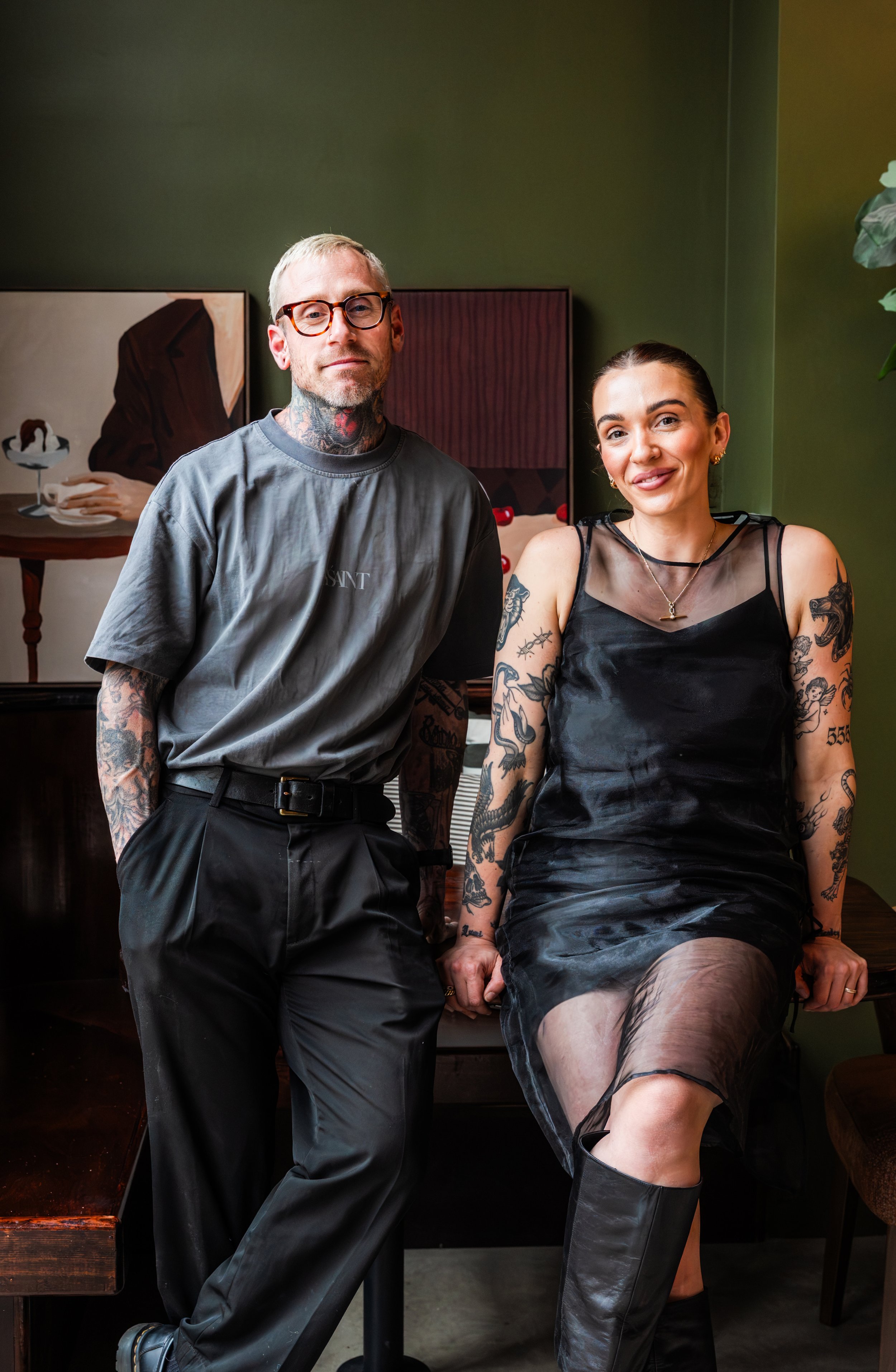 A man with glasses and tattoos standing next to a woman with tattoos sitting on a wooden surface in a room with green walls.
