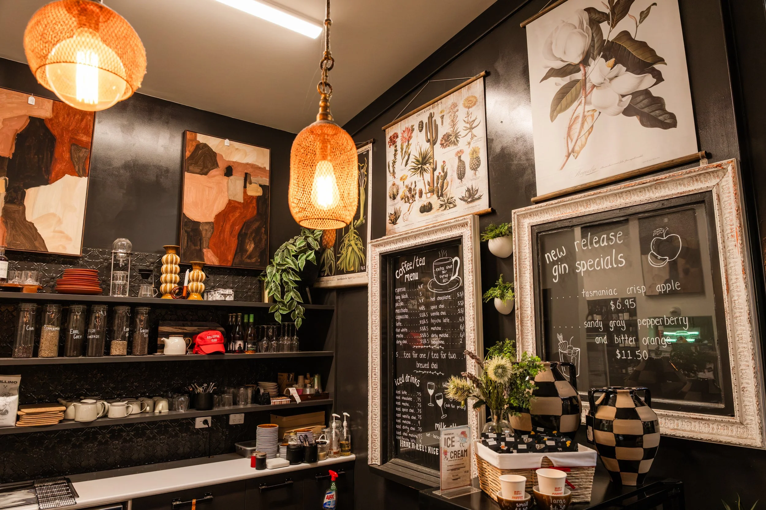 Interior of a cozy coffee shop with black walls decorated with botanical and abstract artwork, warm orange pendant lights, and a chalkboard menu displaying coffee, tea, and new gin specials.