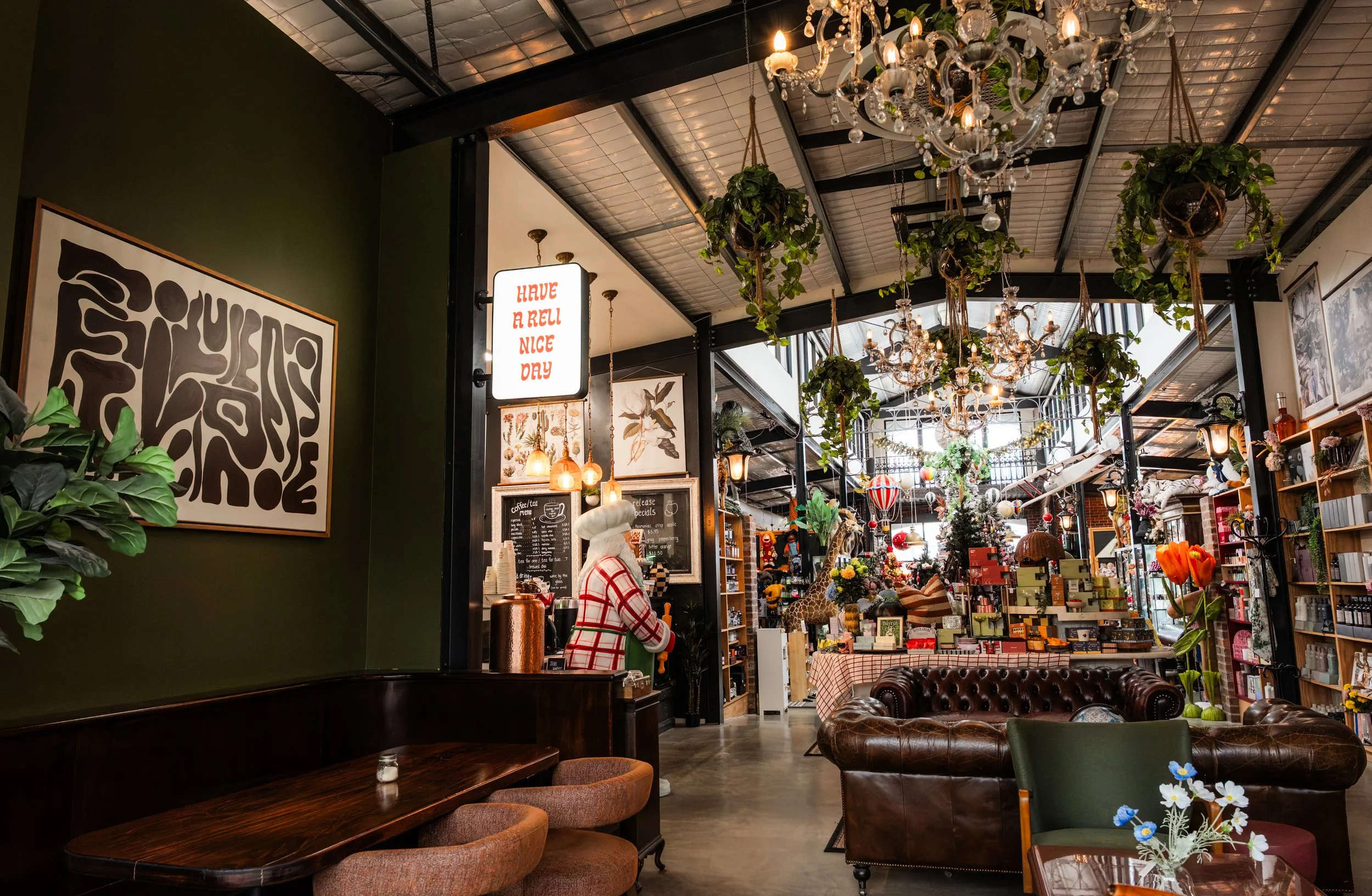 Decorated coffee shop interior with chandeliers, hanging plants, artwork, bookshelves, and plush seating.