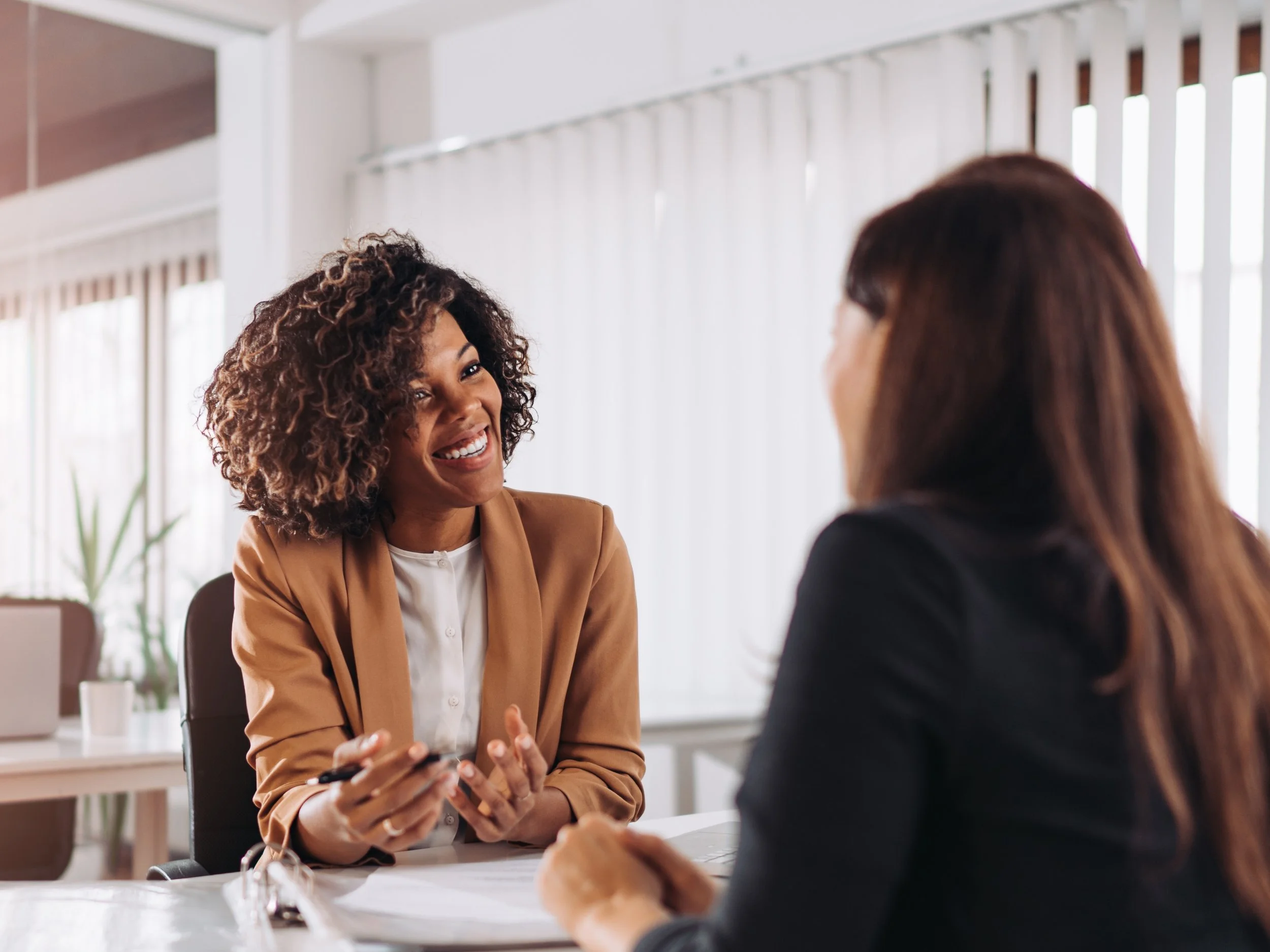 Deux femmes discutent dans un bureau lumineux avec des grandes fenêtres et des rideaux blancs.