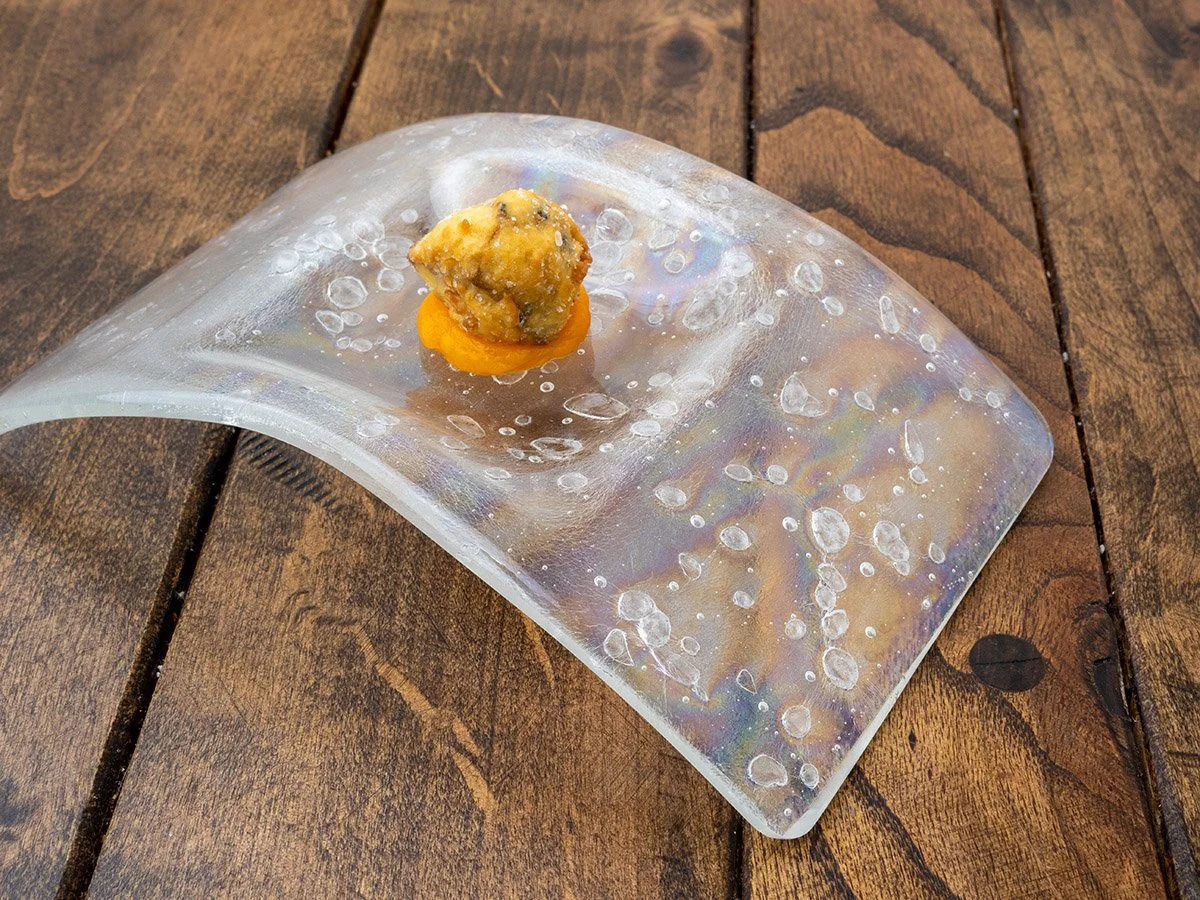 A small piece of fried food on an orange sauce resting on a clear, textured spoon placed on a wooden surface.