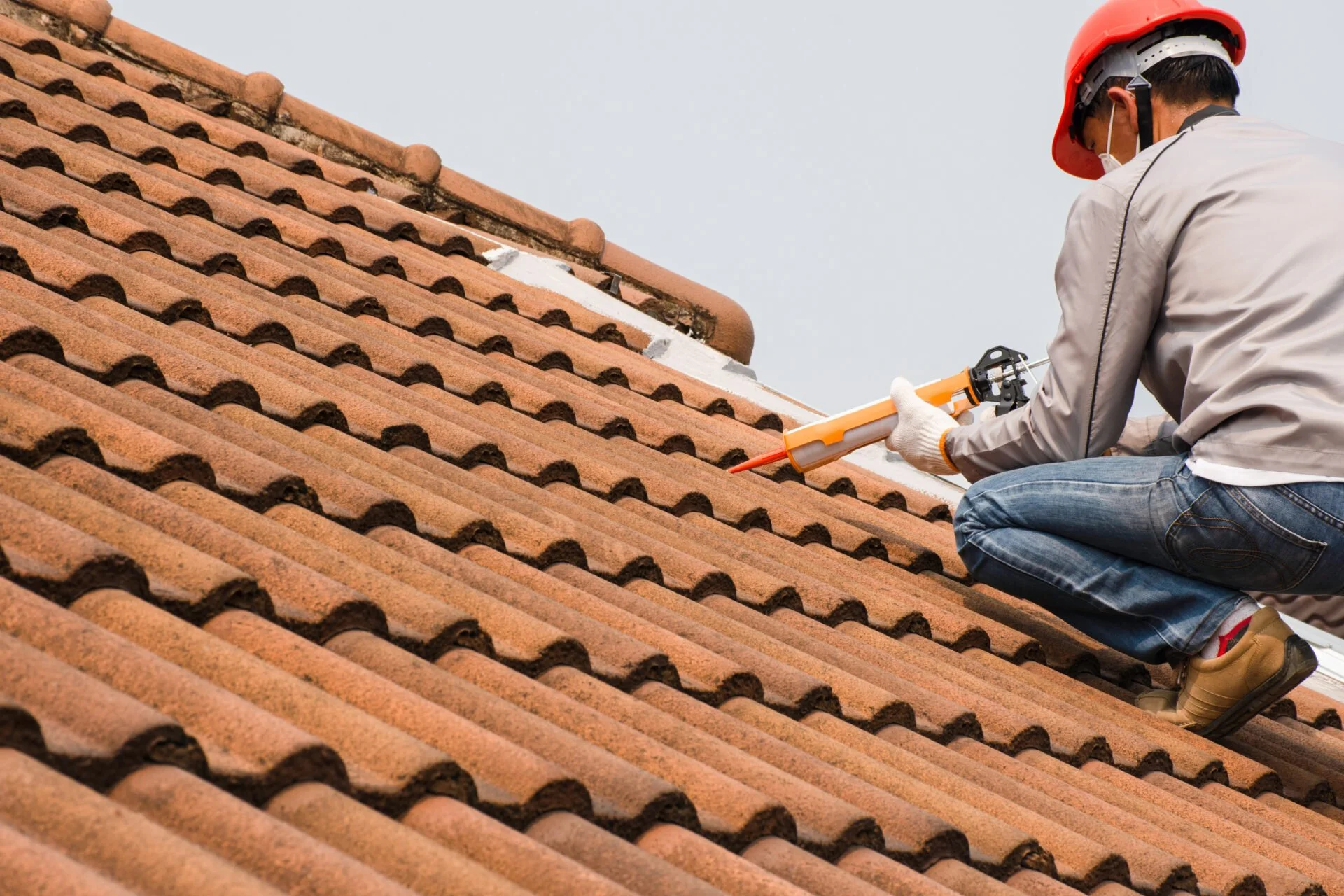 A worker wearing a red safety helmet and beige jacket, kneeling on a tiled roof, applying sealant in a corner with a caulking gun.