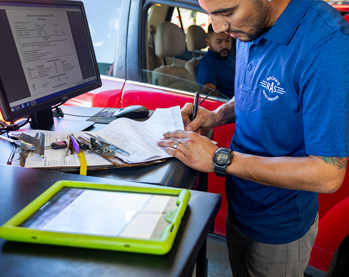Mechanic completing car diagnostic outside a vehicle. There is a computer monitor, a tablet, and various keys on the desk, with a person sitting inside the vehicle in the background.