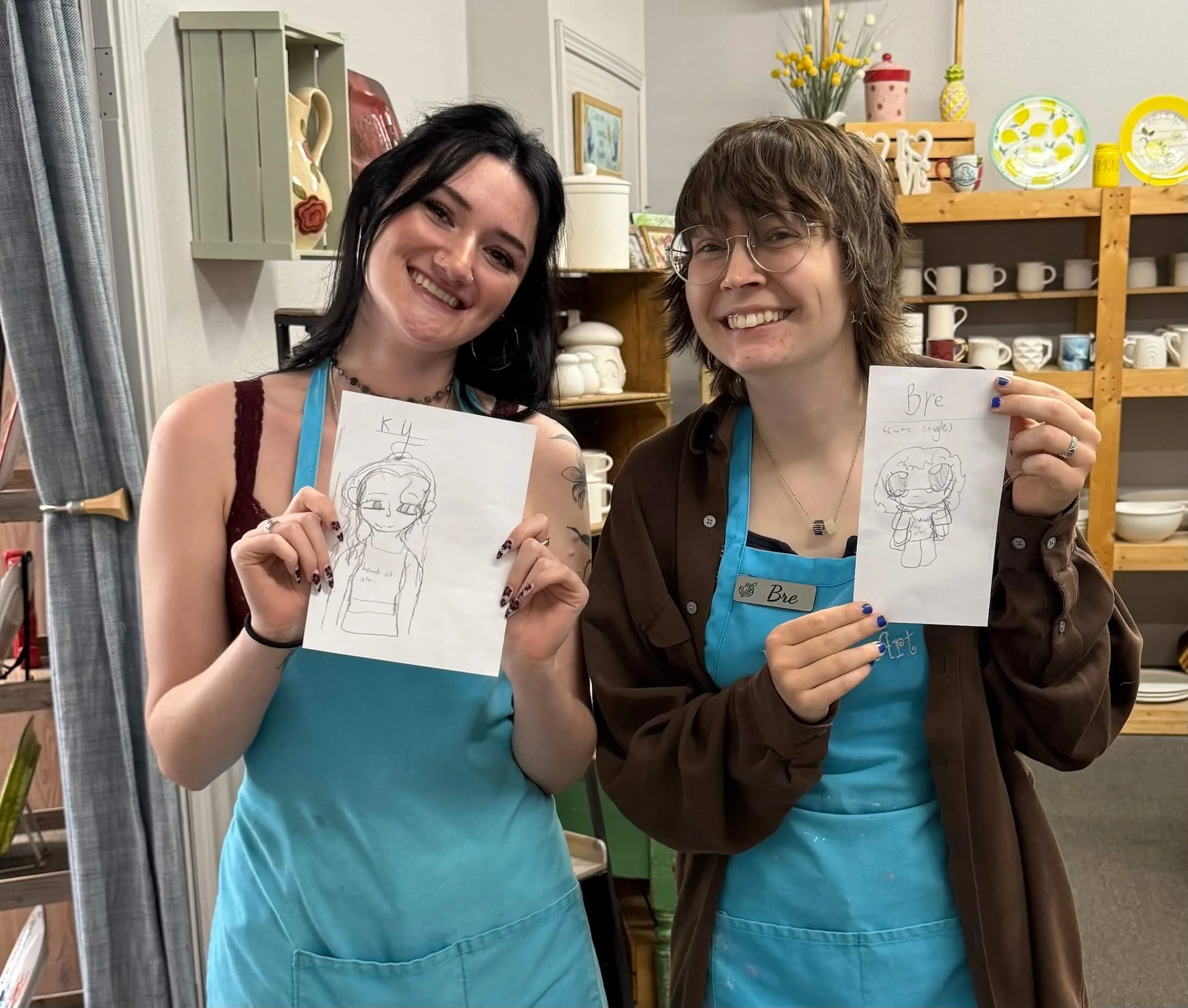 Two people in aqua aprons show off their paper drawings