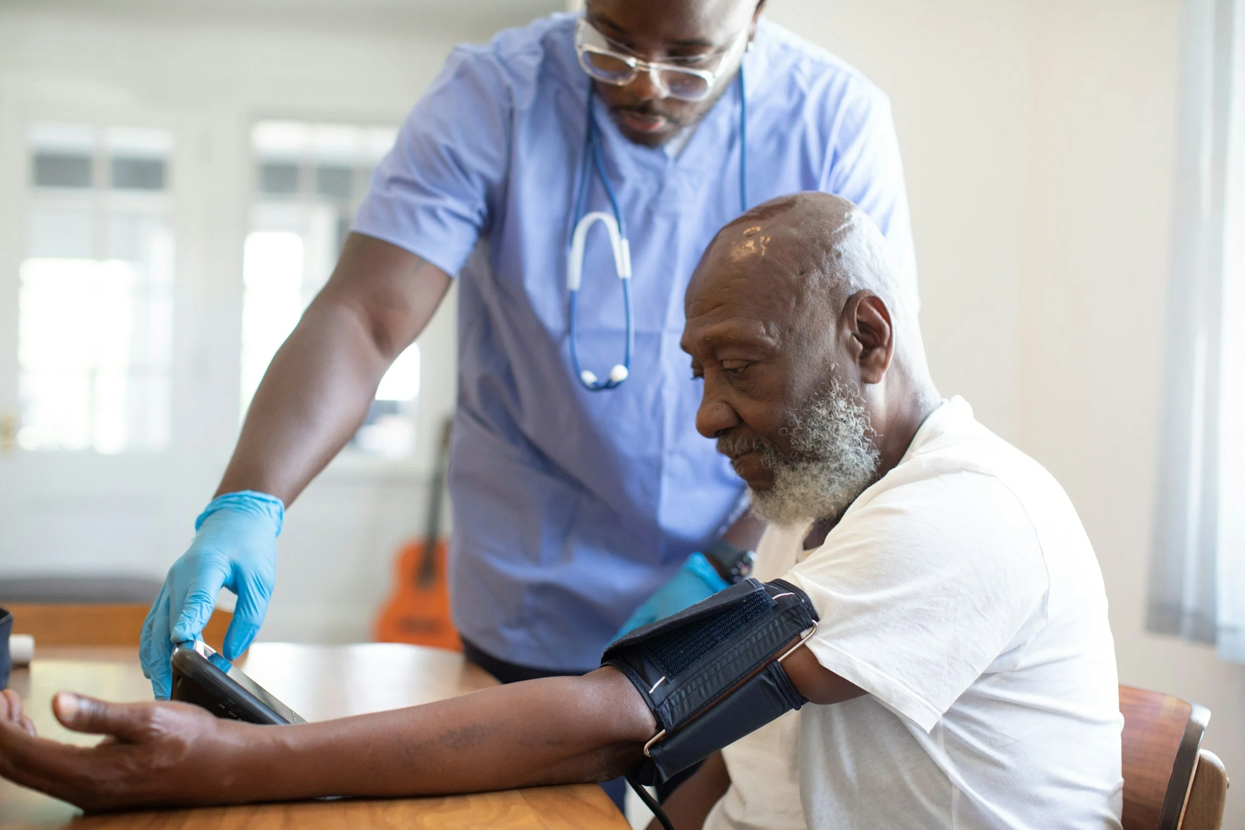 A healthcare professional taking an elderly man's blood pressure with a sphygmomanometer on his arm, using a digital monitor, in a bright room.