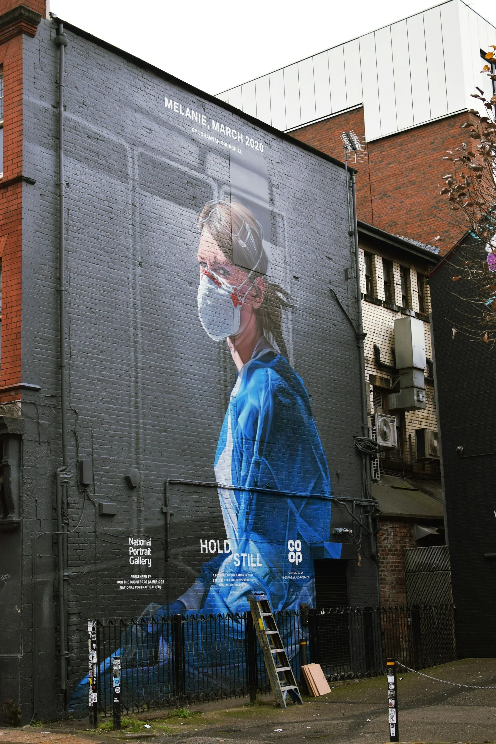 A large mural on a brick building depicts a woman wearing a face mask, with text indicating it was created in March 2020 during the COVID-19 pandemic.