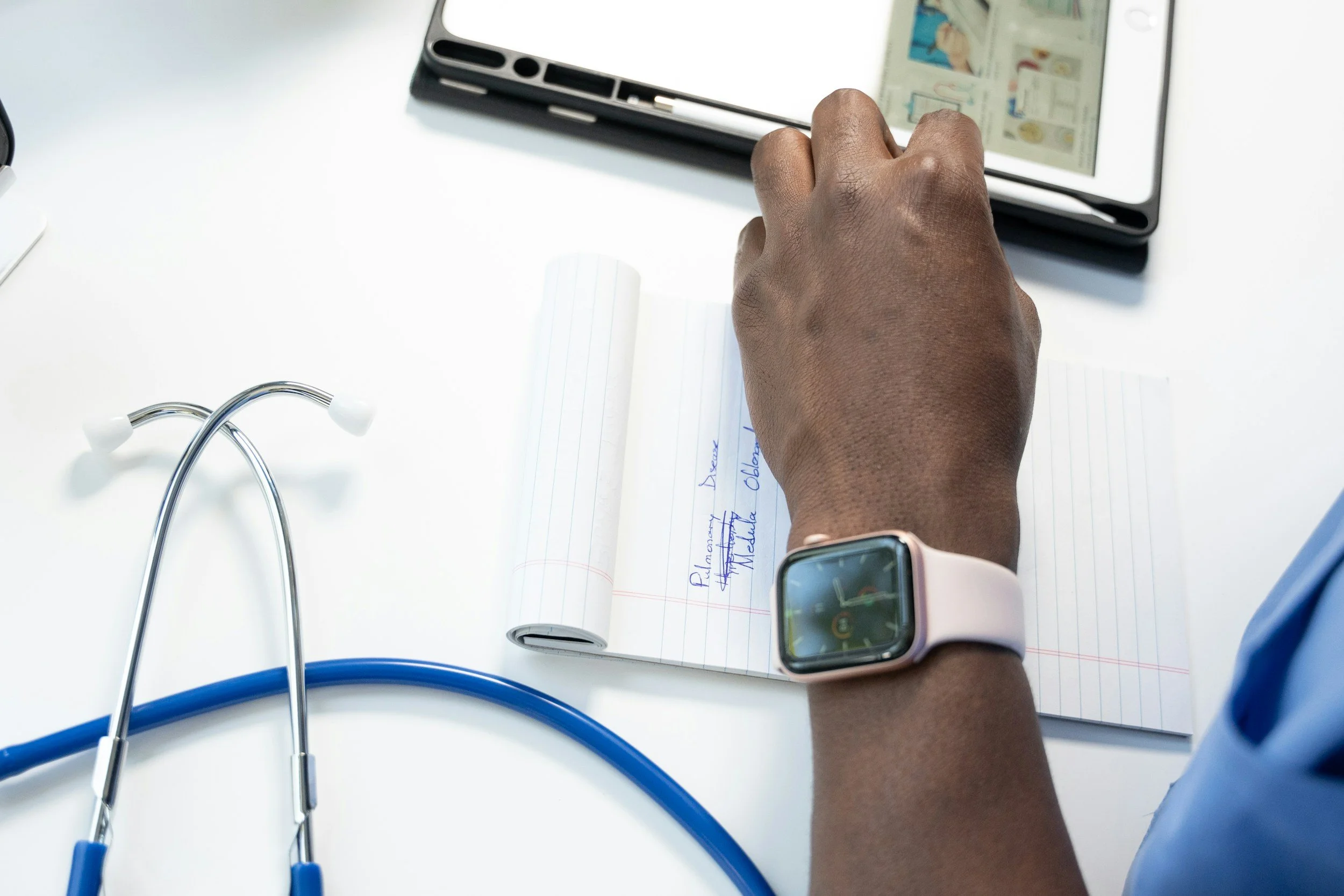 A healthcare professional with a smartwatch on their wrist, writing in a notepad with medical notes, beside a stethoscope on a white desk, and a tablet displaying medical images.
