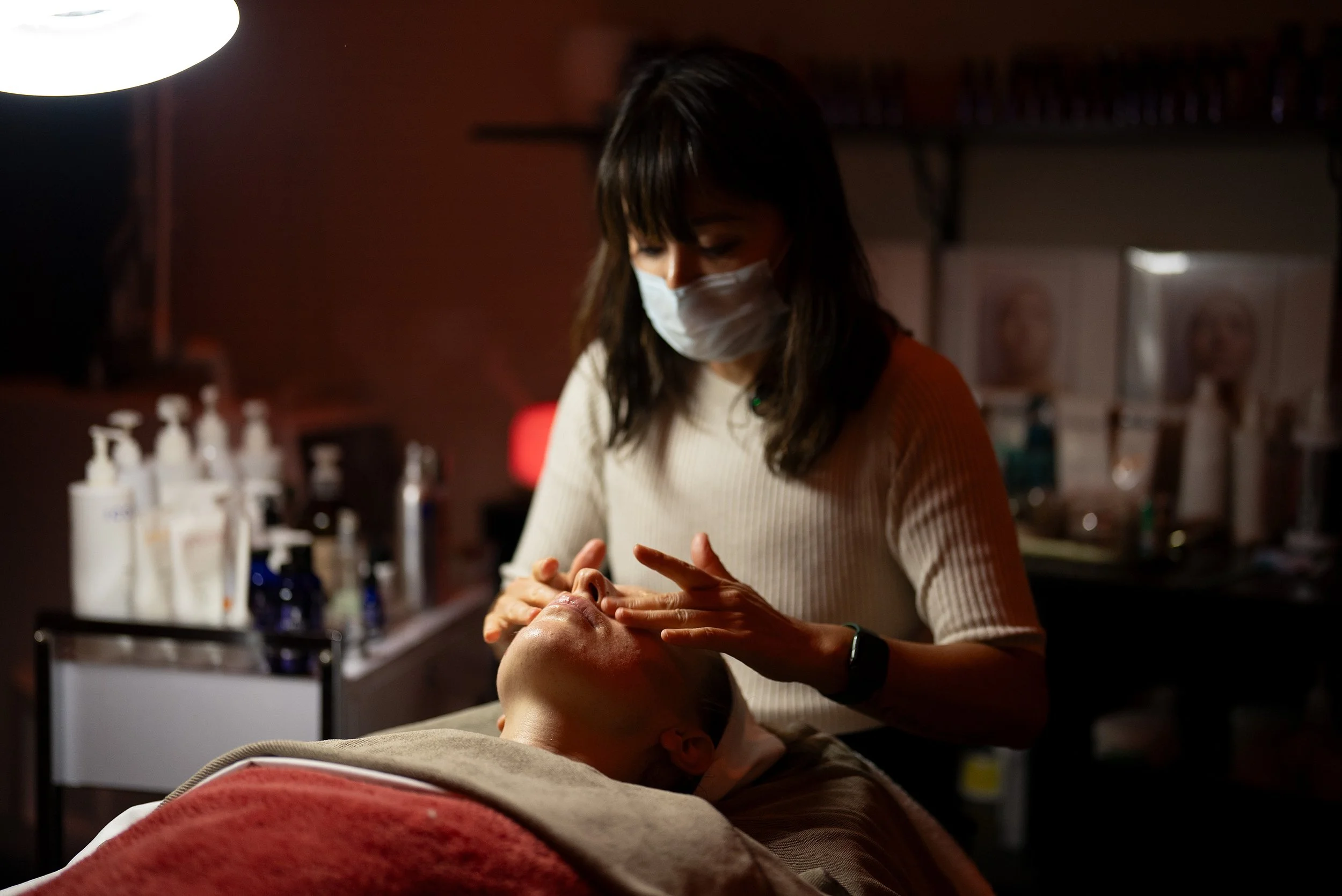 A woman wearing a face mask providing a facial treatment to a client in a dimly lit spa or massage parlor.