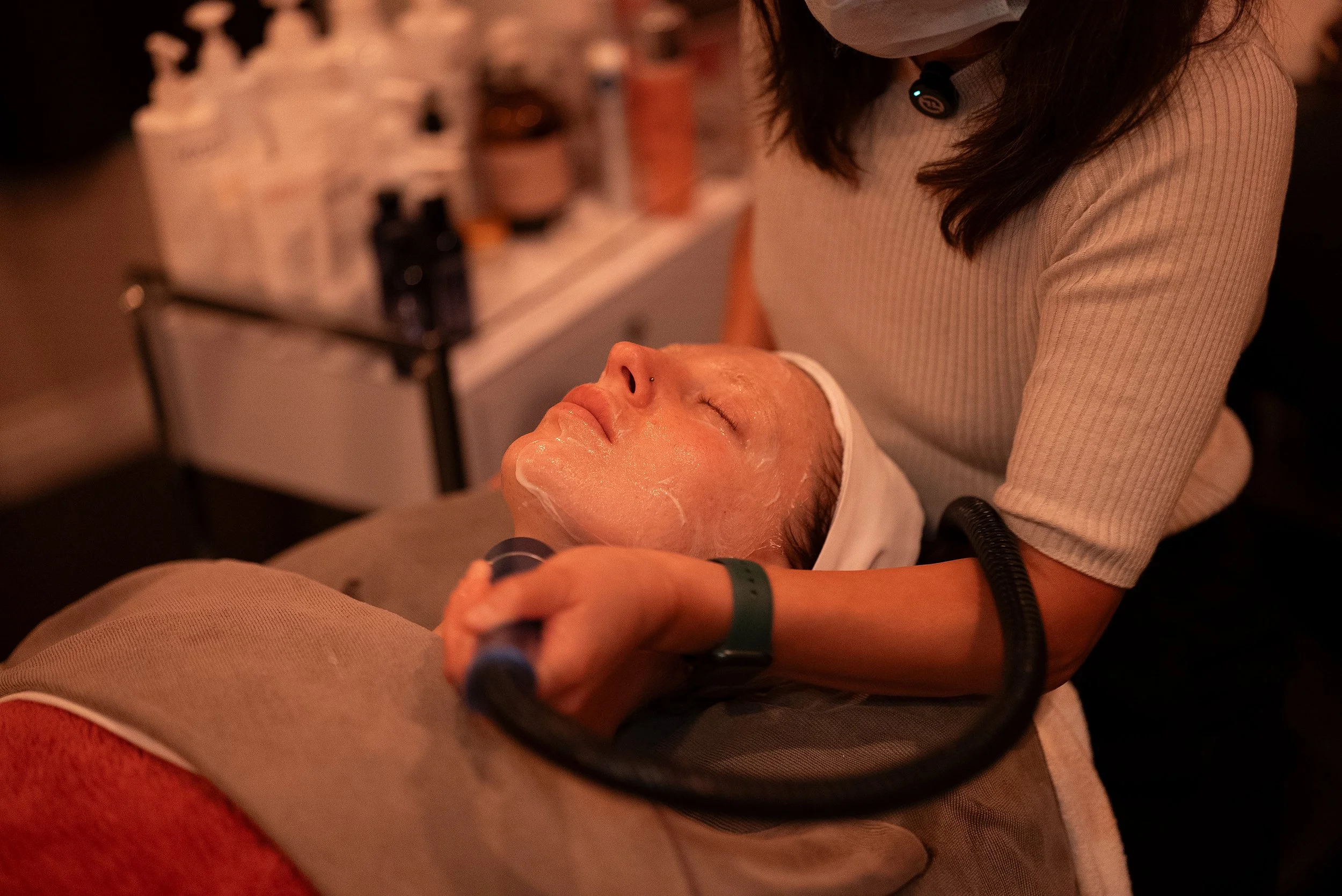 A woman receiving a facial treatment at spa or salon, lying on a treatment bed with her eyes closed, being attended to by a technician in a beige top. The setting is dimly lit, with skincare products and equipment in the background.