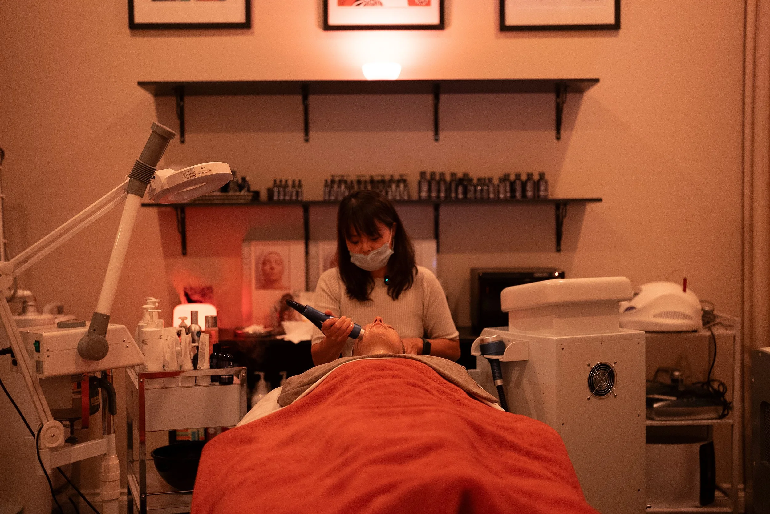 A woman in a mask performing a facial treatment on a patient in a spa or skincare clinic, with skincare products and medical equipment on counters, and shelves with bottles on the wall behind them.
