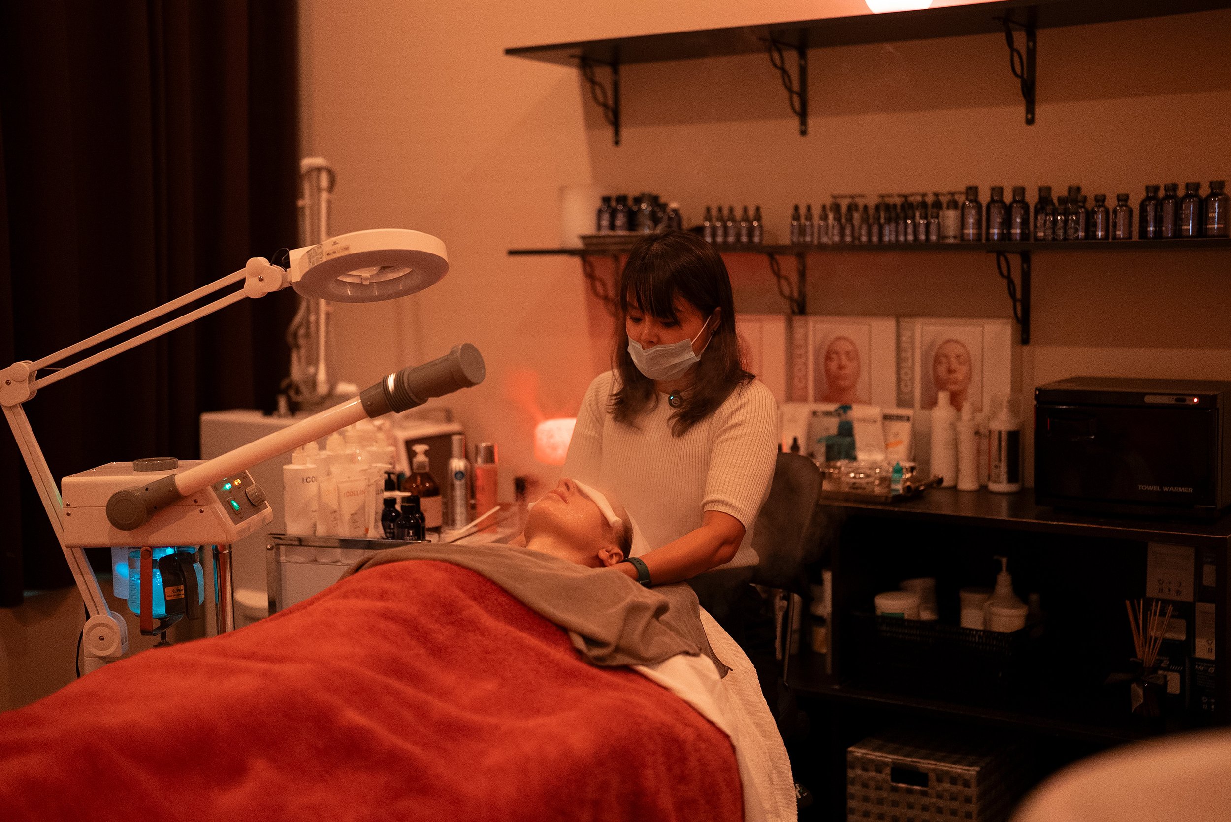 A woman wearing a face mask performing a facial treatment on a patient lying down with a blanket over their body in a skincare clinic. Shelves with bottles and skincare products are in the background.