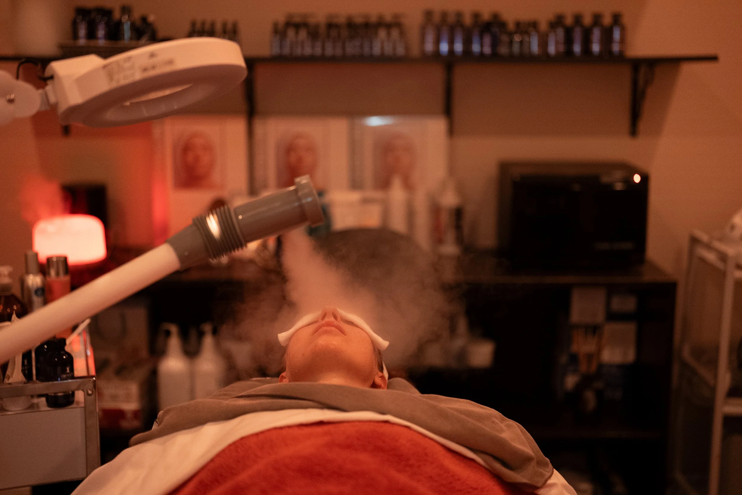 A person lying on a medical bed with a headband, undergoing a facial or skincare treatment in a dimly lit room. Medical equipment and shelves with bottles and jars are visible in the background.