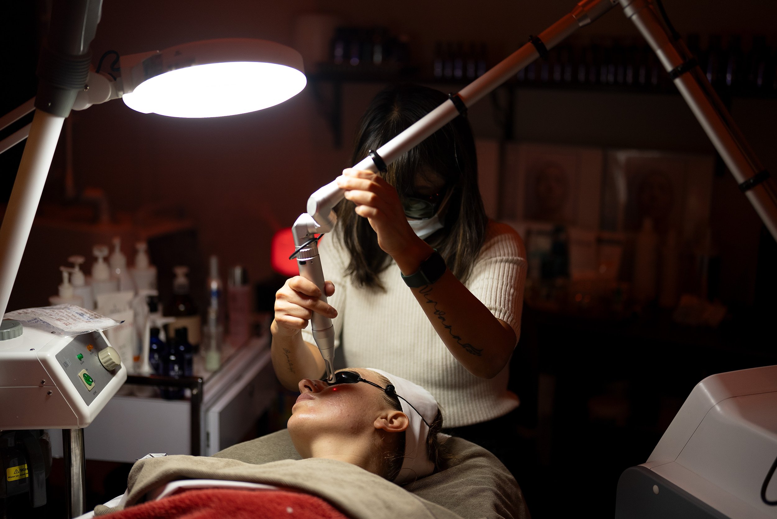 A woman undergoing a laser eye surgery procedure while lying on an operating table in a clinical setting with medical equipment. A technician is using a handheld laser device near her eye under a bright operating light.