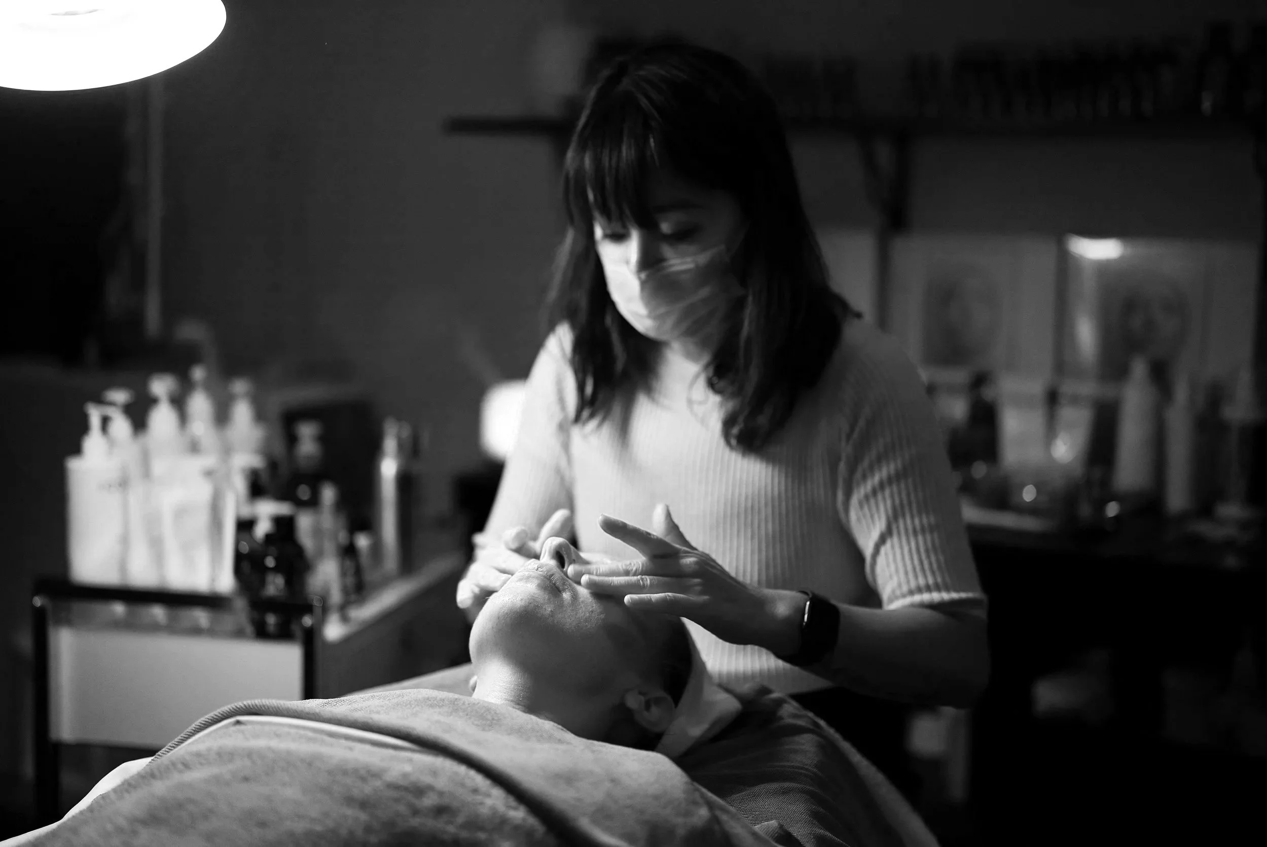A woman wearing a face mask performs a facial treatment on a client lying down in a spa or salon setting.