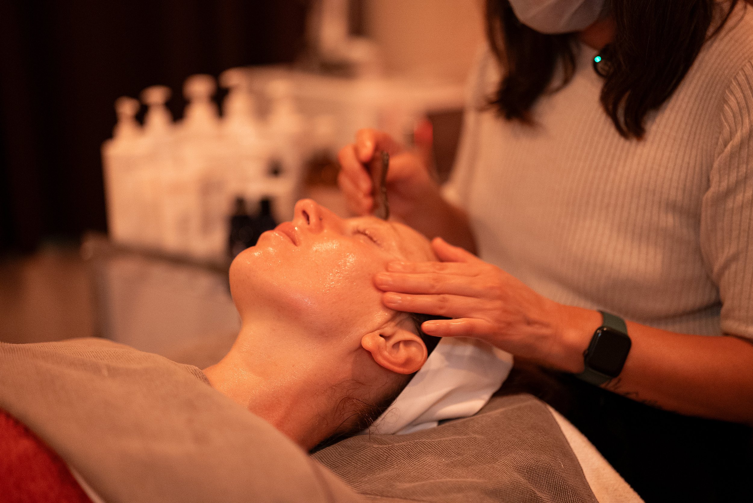 A person is receiving a facial treatment or skincare application in a spa or wellness setting, with the therapist or skincare specialist gently working on the client's face.