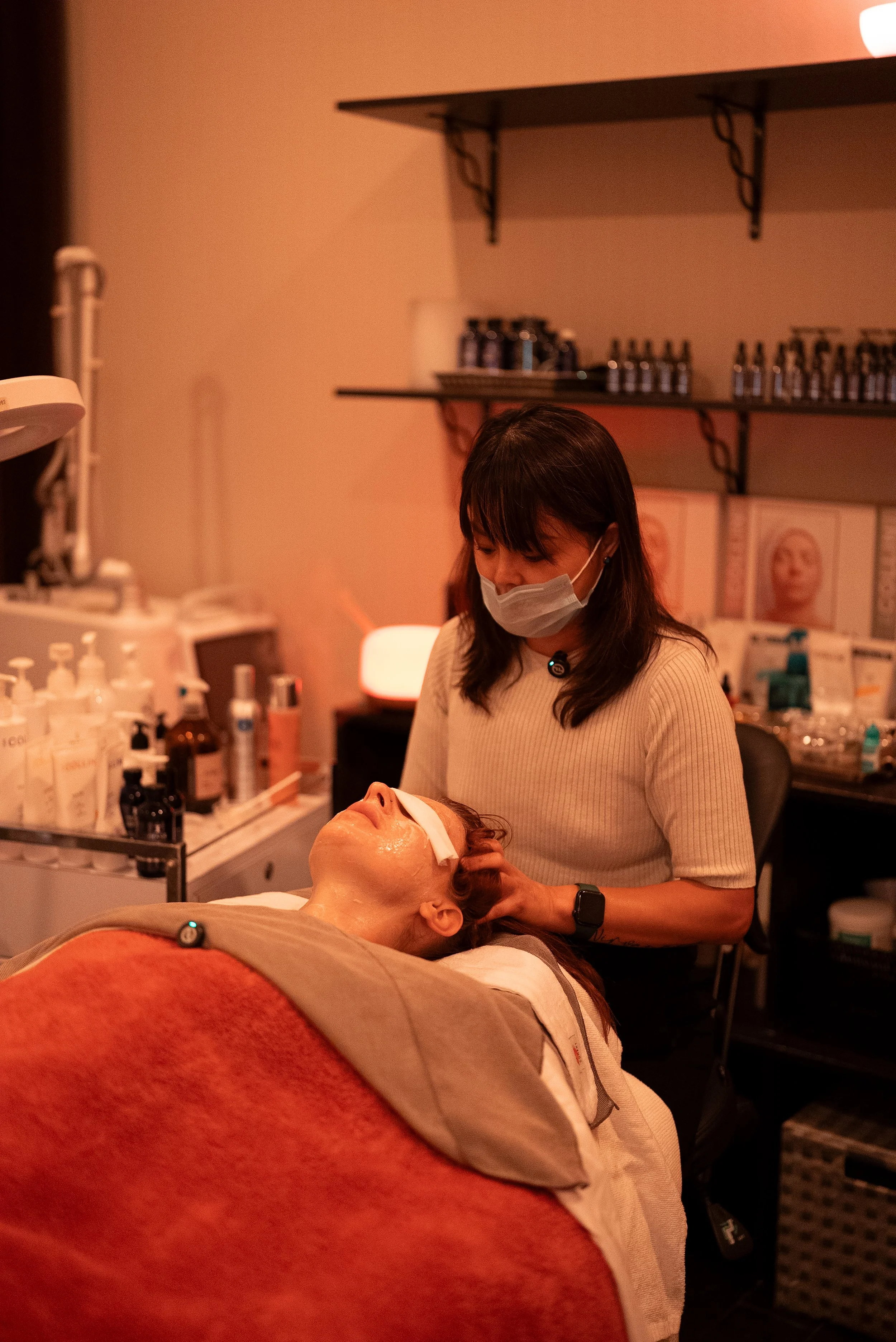 A woman receiving facial treatment from an esthetician in a spa or clinic with shelves of skincare products in the background.