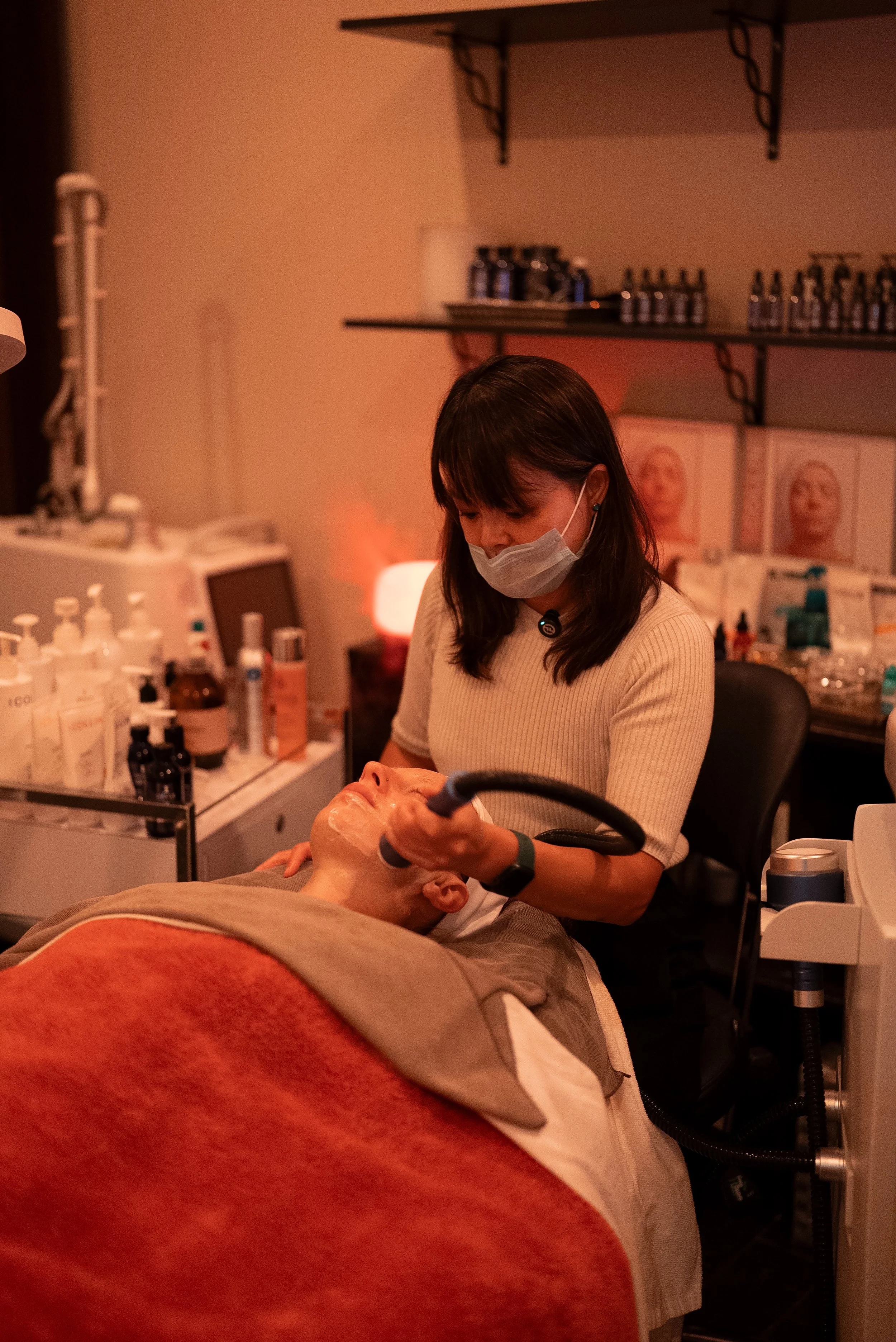 A woman wearing a mask is performing a facial treatment on a client lying on a bed. The room has medical supplies and skincare products on nearby tables, and shelves with bottles and boxes in the background.