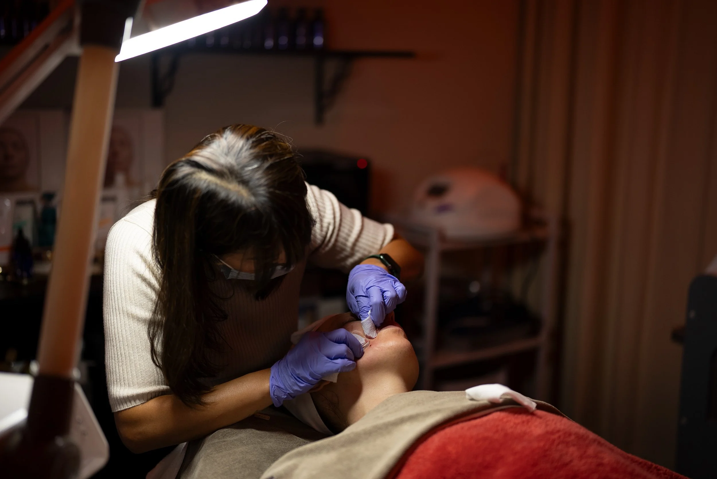 A person with long hair receiving a tattoo on their body from a tattoo artist in a studio. The tattoo artist is wearing gloves and working under a bright light.