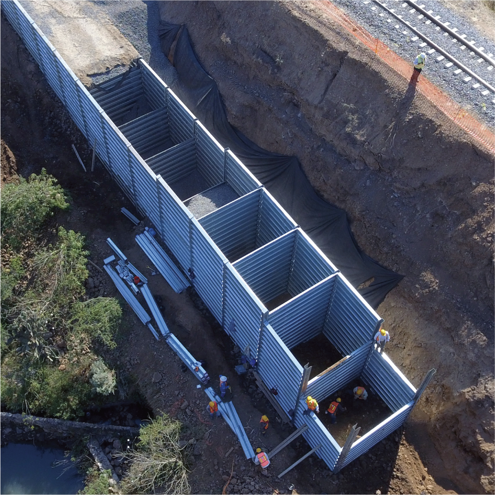 Construcción en progreso de una estructura de paneles de metal en un socavón, con trabajadores en casco y equipo de seguridad, junto a vías del tren.