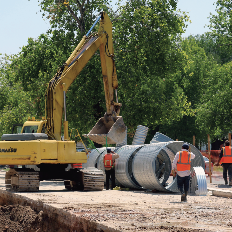 Maquinaria de construcción y trabajadores en un sitio de obras, manipulando tuberías metálicas en un entorno exterior con árboles verdes.