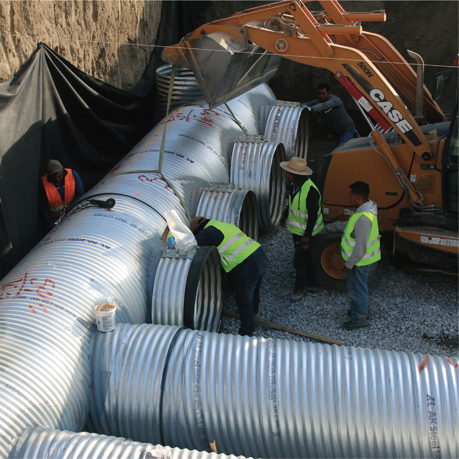 Trabajadores en una excavación colocando grandes tuberías de acero con ayuda de una máquina excavadora.