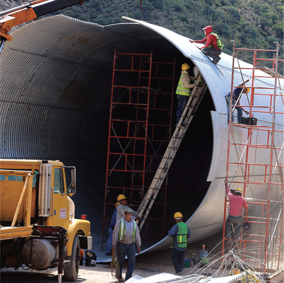 Trabajadores ensamblando o inspeccionando una estructura metálica grande en un área al aire libre con colinas verdes al fondo, usando equipo de protección y andamios.