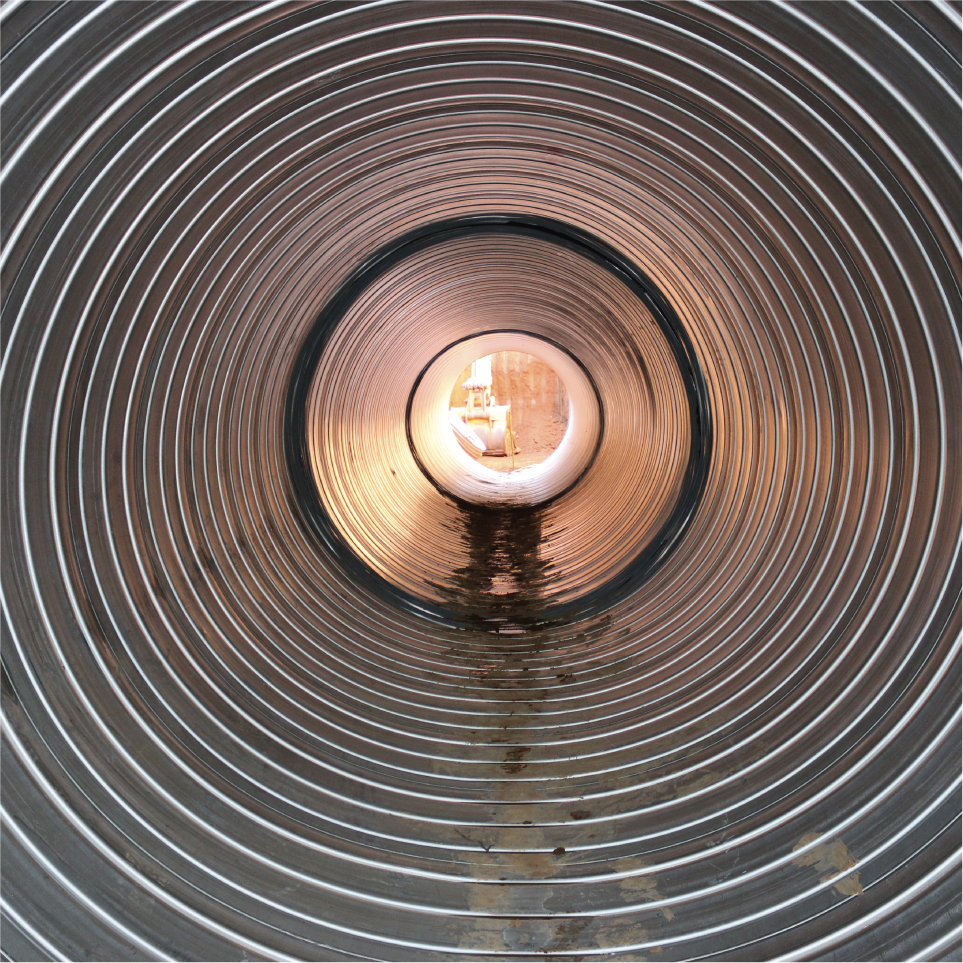 Vista desde dentro de un tubo de metal con superficie ondulada, con luz brillante en el extremo opuesto y charcos de agua en el interior.