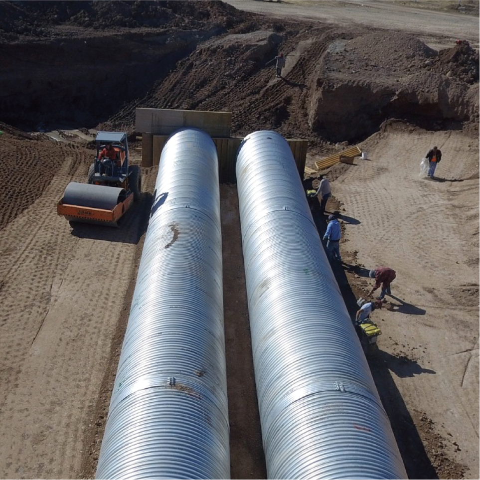 Construcción de tuberías metálicas en un sitio de obra, con maquinaria y personas trabajando en el terreno.
