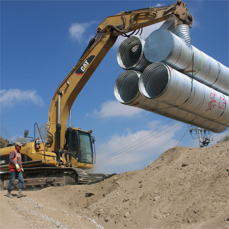 Excavadora amarilla colocando tuberías de metal en un sitio de construcción bajo cielo despejado.