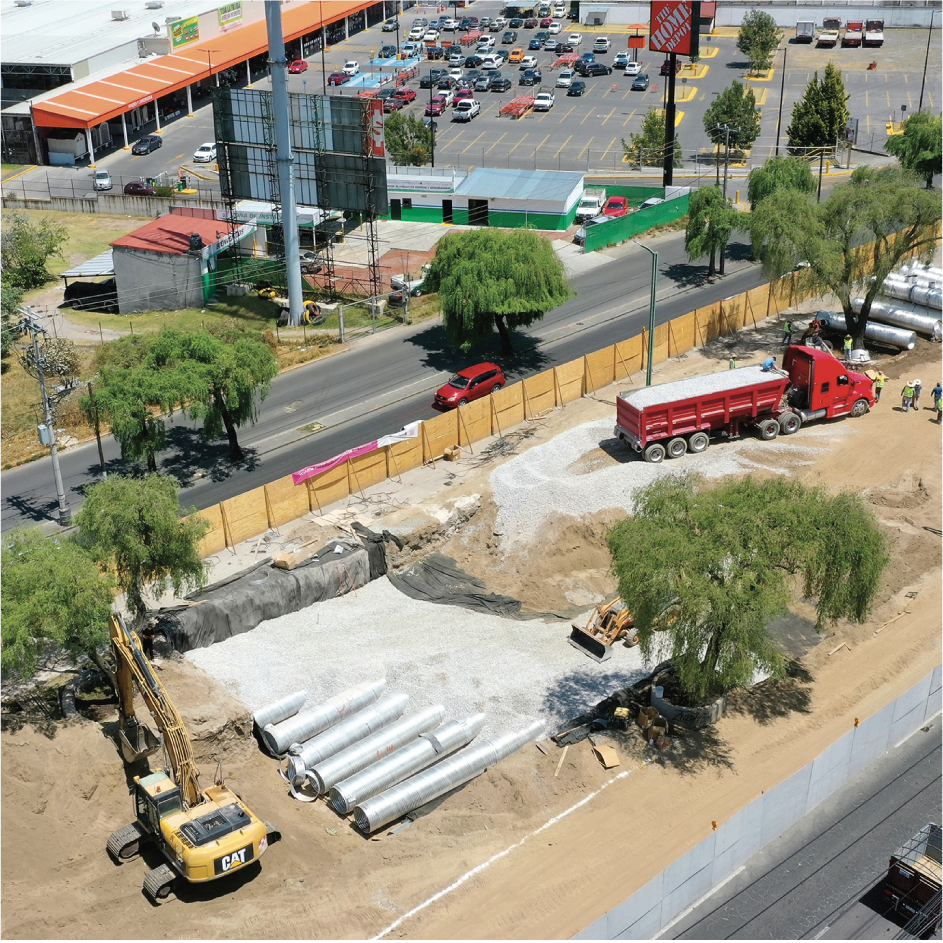 Construcción de un proyecto de infraestructura con maquinaria pesada, tuberías de metal y una cerca de madera, en un área en construcción junto a la calle, con tráfico y un estacionamiento en el fondo.