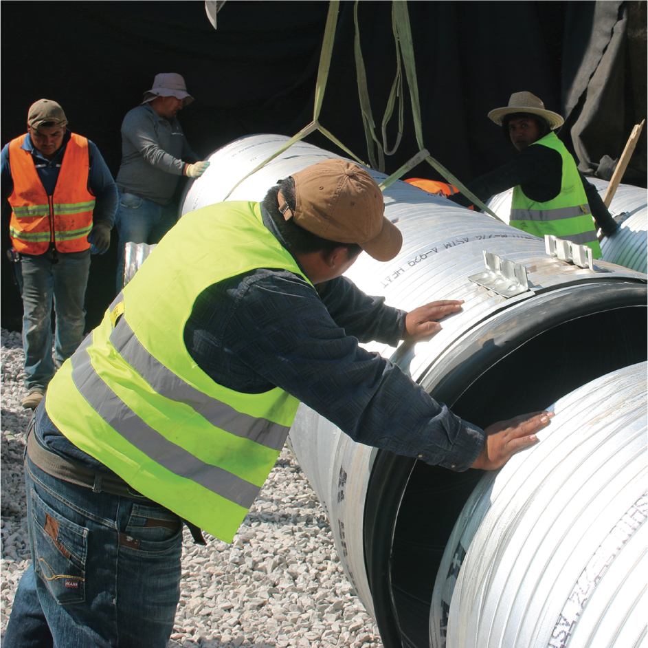 Trabajadores instalando tuberías grandes en una obra de construcción, usando chalecos de seguridad y guantes.