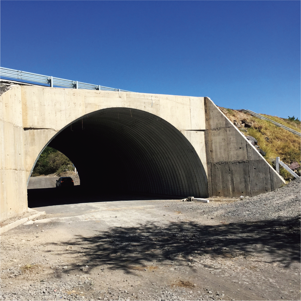 Puente de concreto con forma de arco sobre una carretera en un área rural, con un cielo despejado y colinas al fondo.