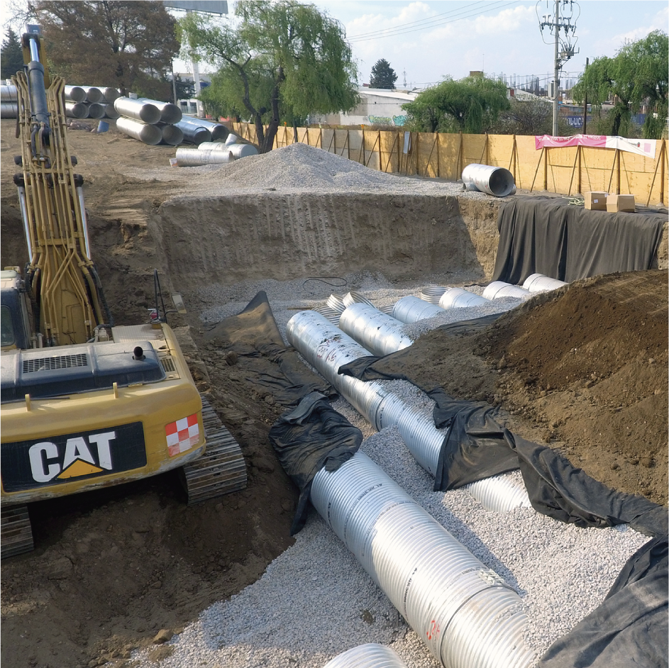 Equipos de construcción y tuberías de metal en una obra en construcción, con máquinas excavadoras y una cerca de madera, en un entorno al aire libre con árboles y estructuras urbanas en el fondo.