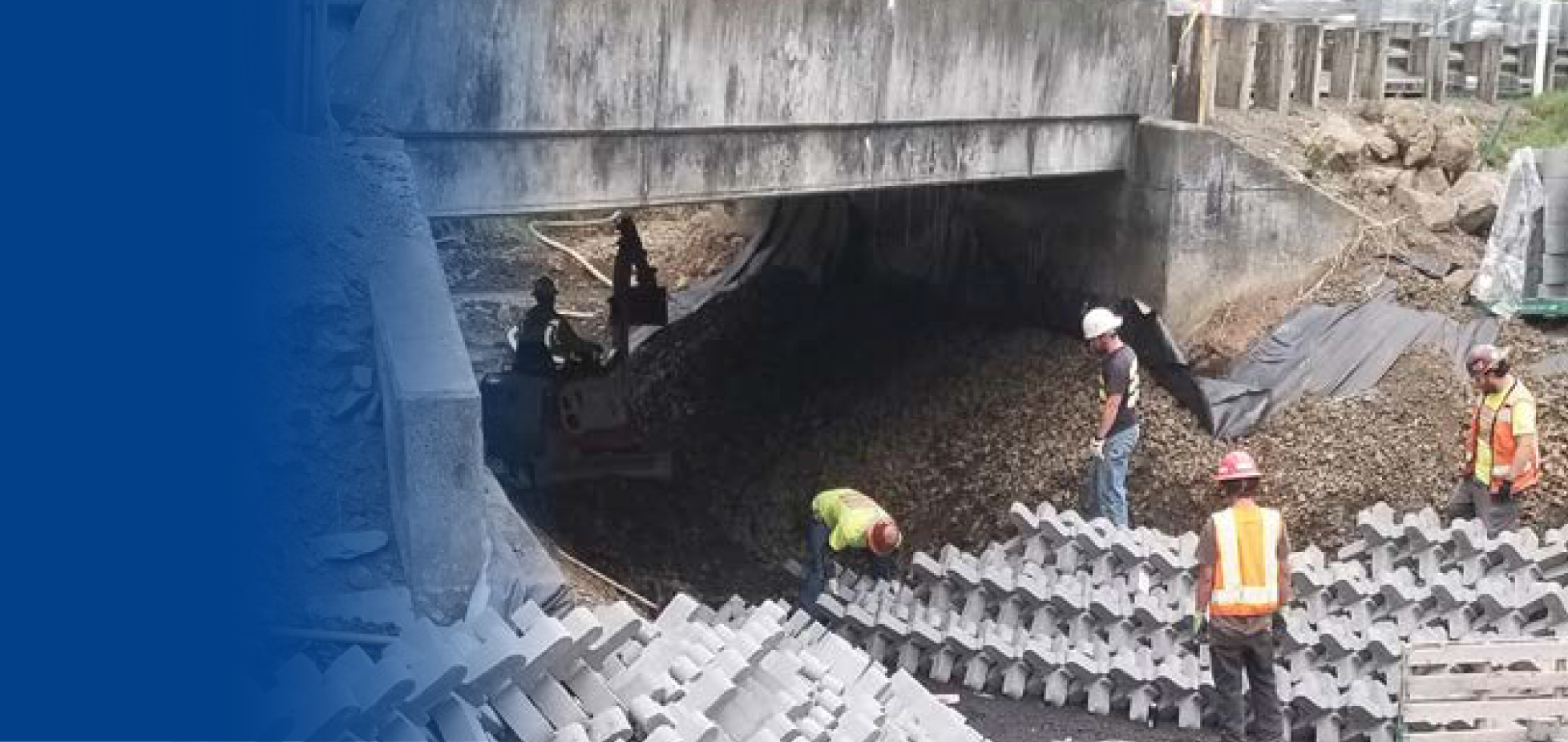 Trabajadores de construcción en un sitio de obras civiles debajo de un puente, colocando bloques de concreto en la estructura.