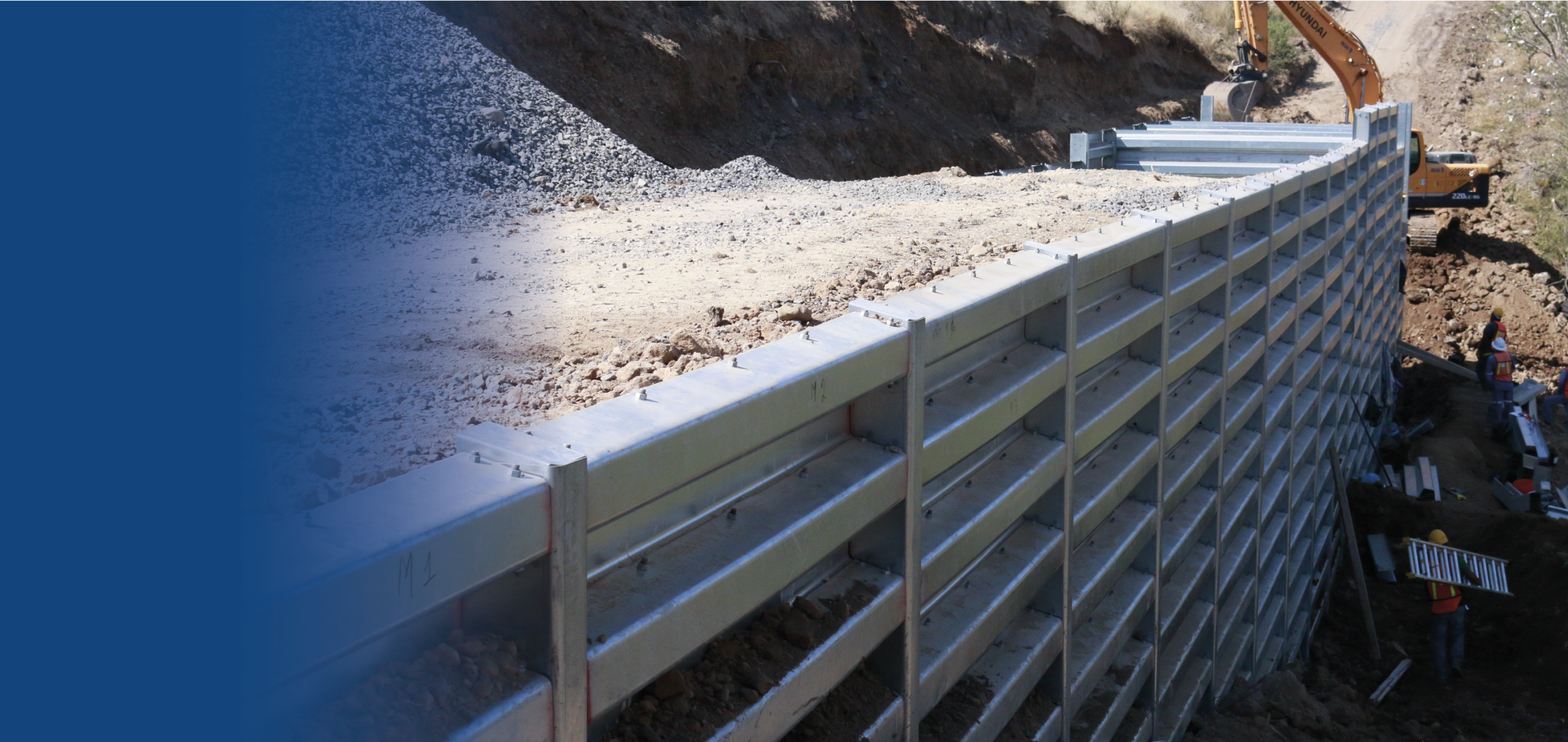 Construcción de muro de contención de metal en un proyecto de obras civiles en un terreno montañoso, con maquinaria y trabajadores.