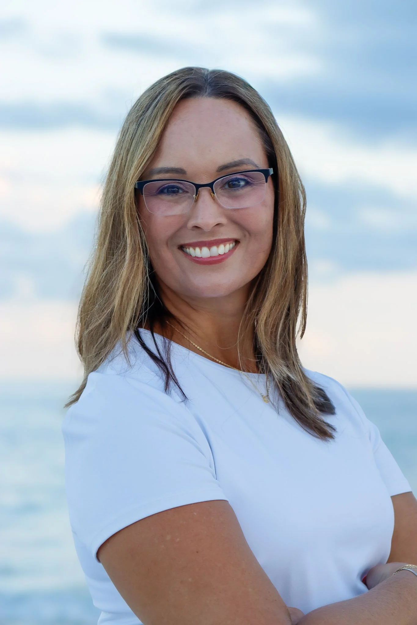 A woman, counselor/therapist, with light brown hair, glasses, and a white shirt smiling outdoors against a background of the ocean and sky.