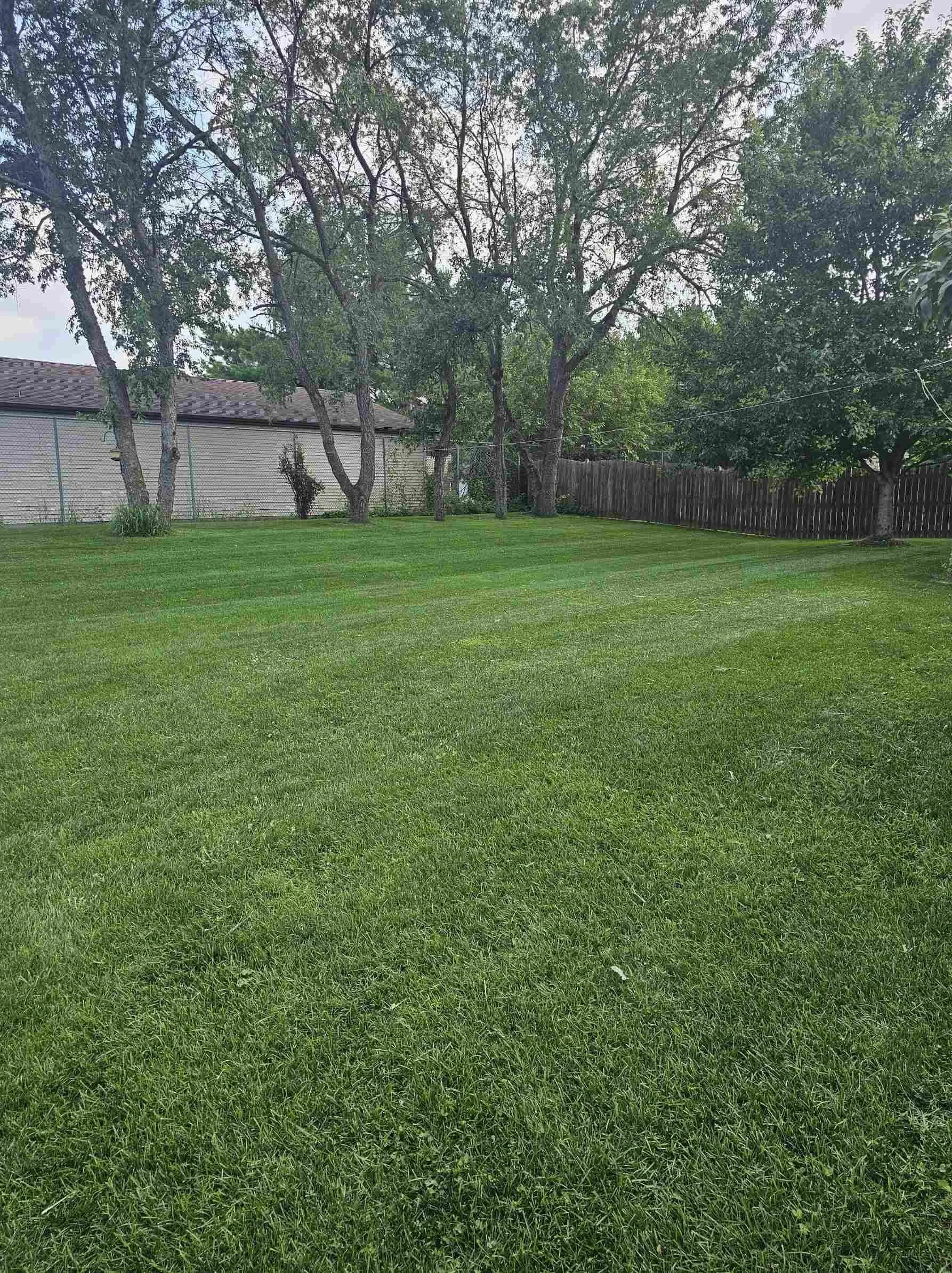 A backyard with well-maintained green grass, trees along the fence, and a shed or house with a brick exterior in the background.