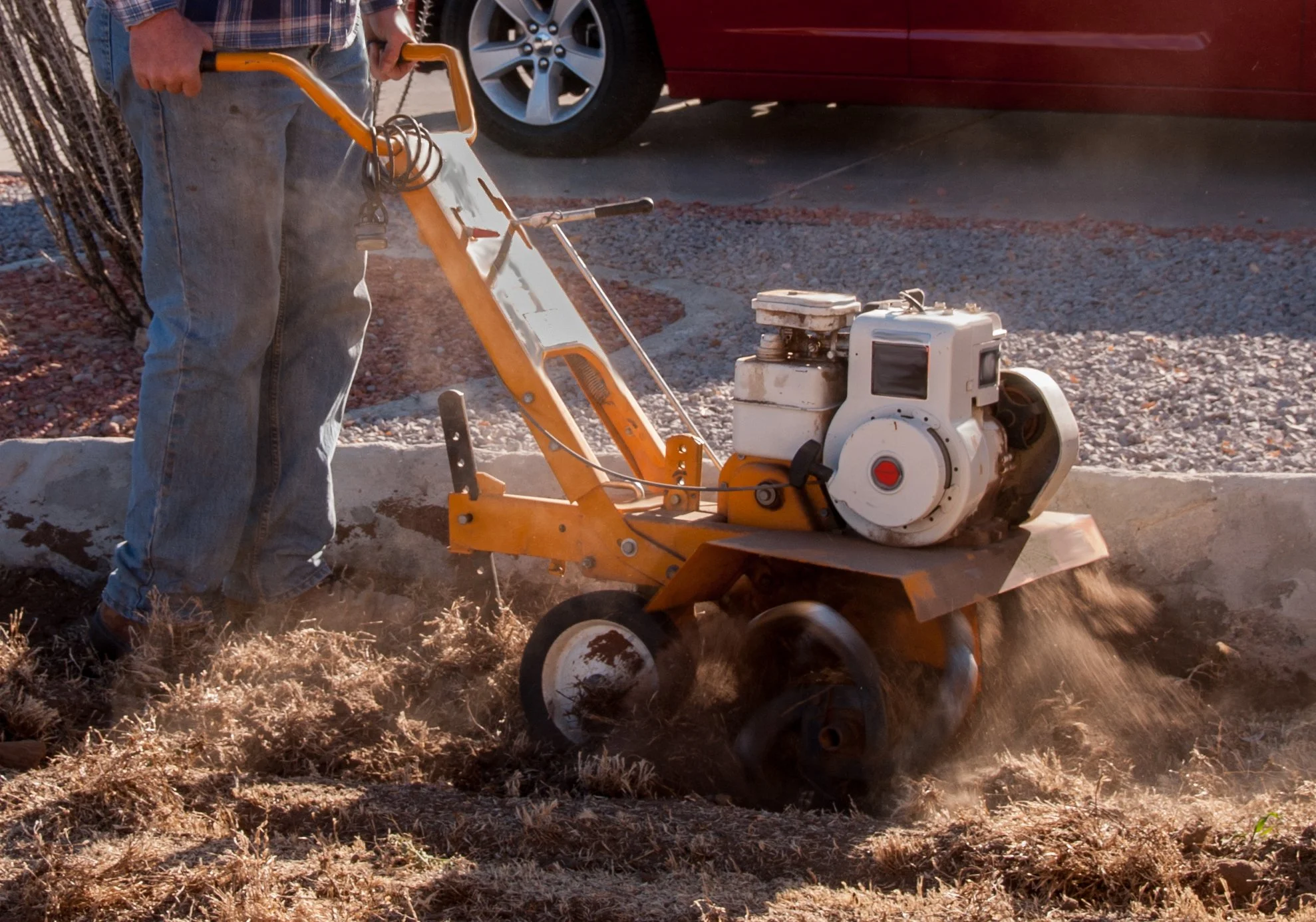 Person pushing a vibratory plate compactor on dirt, with car and gravel in background.